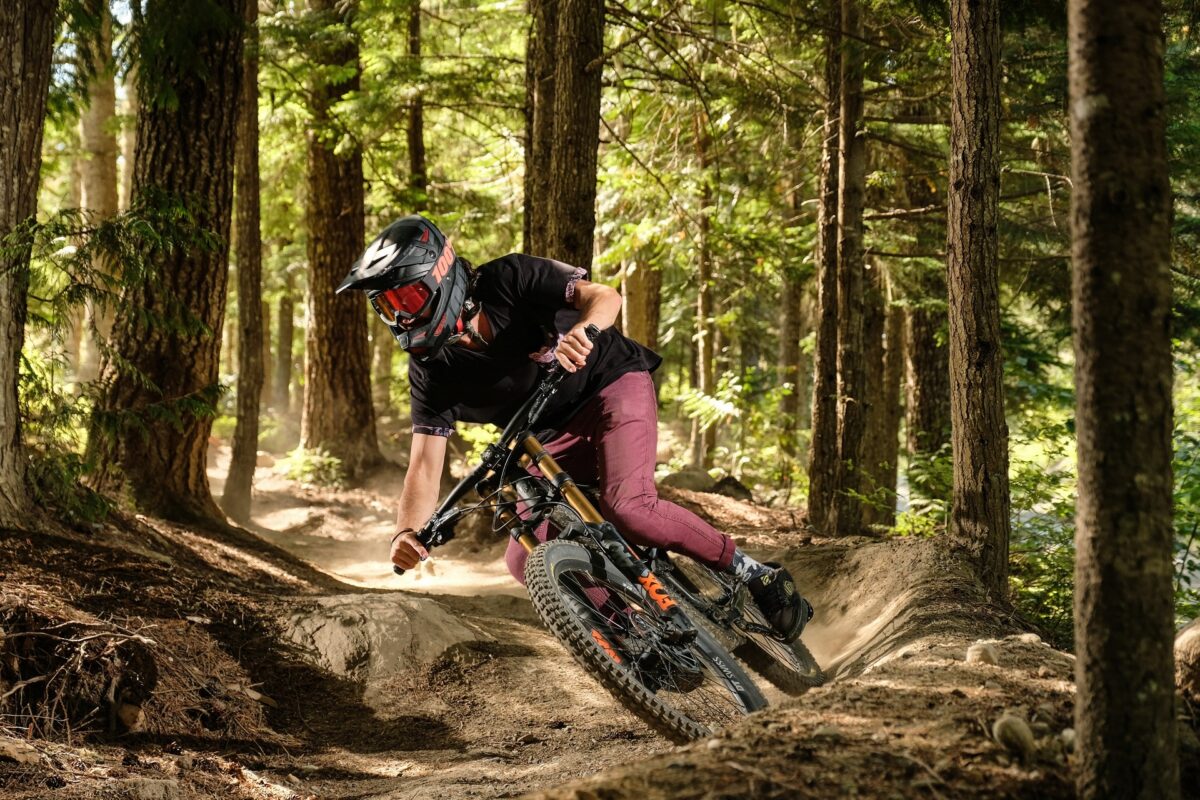 A mountain biker is riding his bike in the forest. It is sunny and warm. He is wearing a helmet and leaning into the curve at full speed.