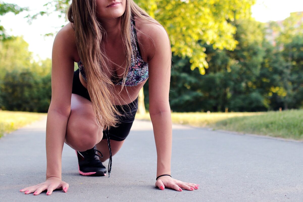 A young woman stretches on the sidewalk of a park. It's sunny and she's looking forward to working on her athletic performance again after her break from training.